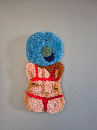 Vægtæppet Mona er en del af en badenymfeserie, der hylder kropspositivitet, farveglæde og det gode humør. Værket viser en rygvendt figur i en klar rød bikini under en stor, lyseblå solhat, hvilket giver et herligt, sommerligt udtryk.